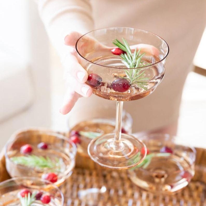 Person holding acrylic champagne glass filled with festive cocktail garnished with rosemary and cranberries.