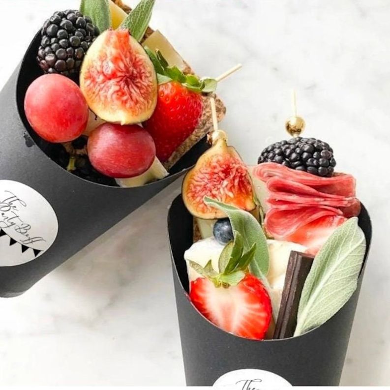 Black charcuterie cup filled with fresh fruit, cheese, and meats, styled with gold fancy toothpick.