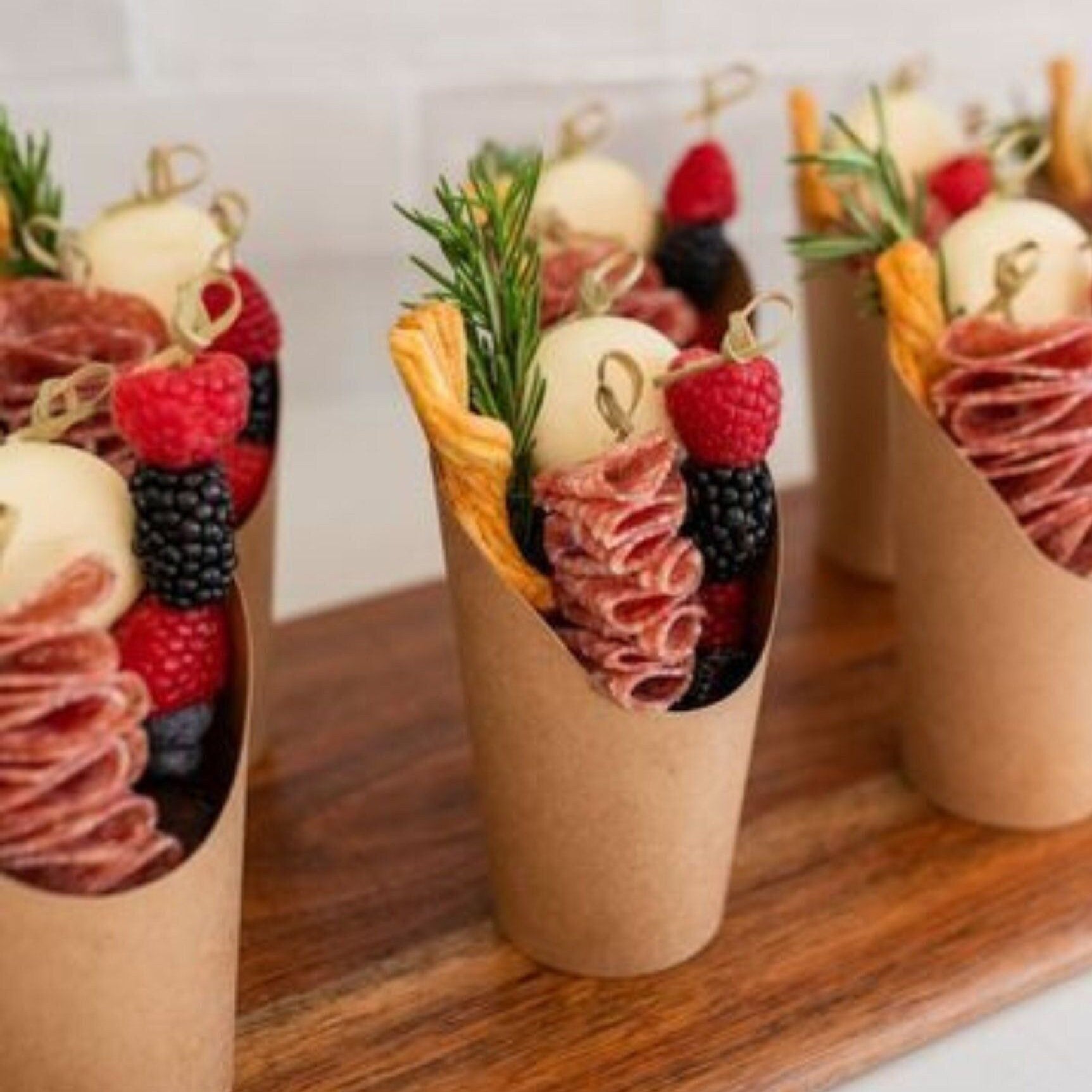 Brown kraft charcuterie cup filled with meats, cheese, and fruit, paired with a fancy wooden toothpick.