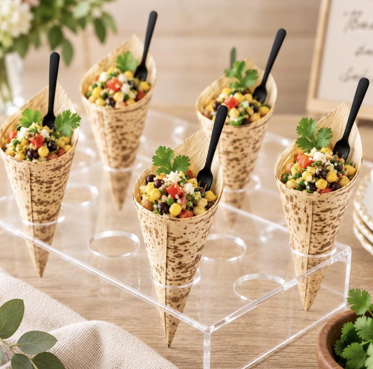 Bamboo appetizer cones with black mini forks filled with corn, beans, tomatoes and fresh herbs arranged in a clear catering display stand