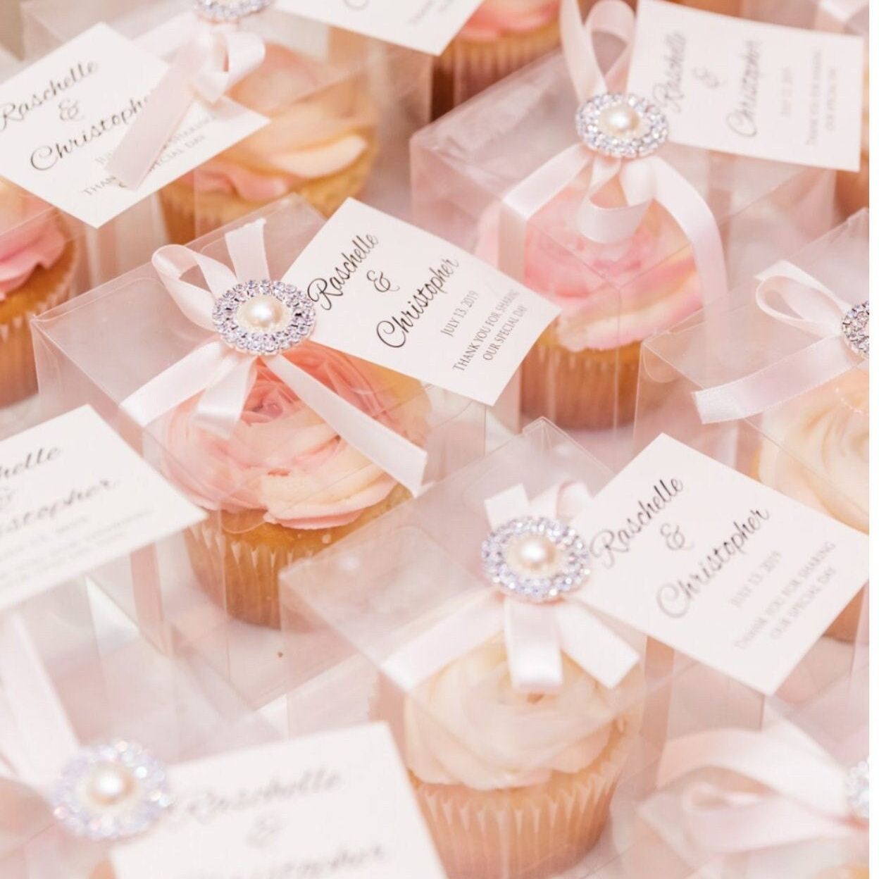 Multiple clear cupcake boxes arranged with decorative ribbons and gift tags for special occasions. Transparent favor boxes hold beautifully decorated cupcakes perfect for bridal showers and celebrations.