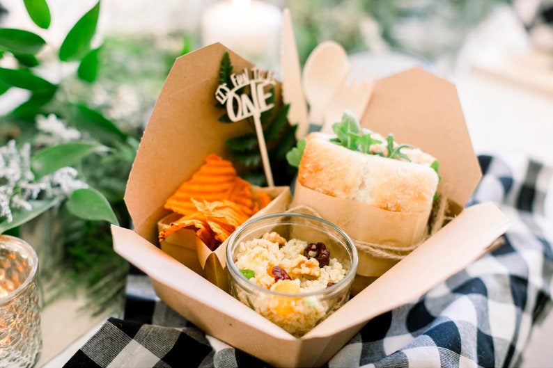 Kraft paper lunch box filled with charcuterie meats, fresh salad, and gourmet sandwich at wedding reception table.