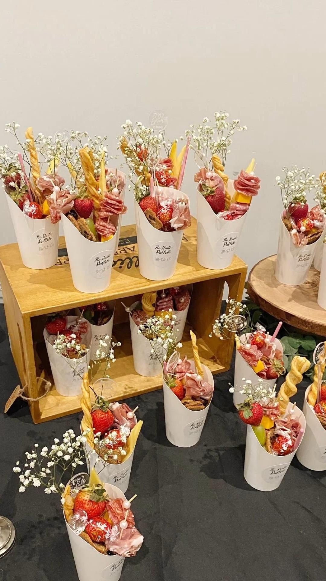 Multiple white charcuterie cups arranged on black table with assorted grazing appetizers and gold fancy toothpicks display.