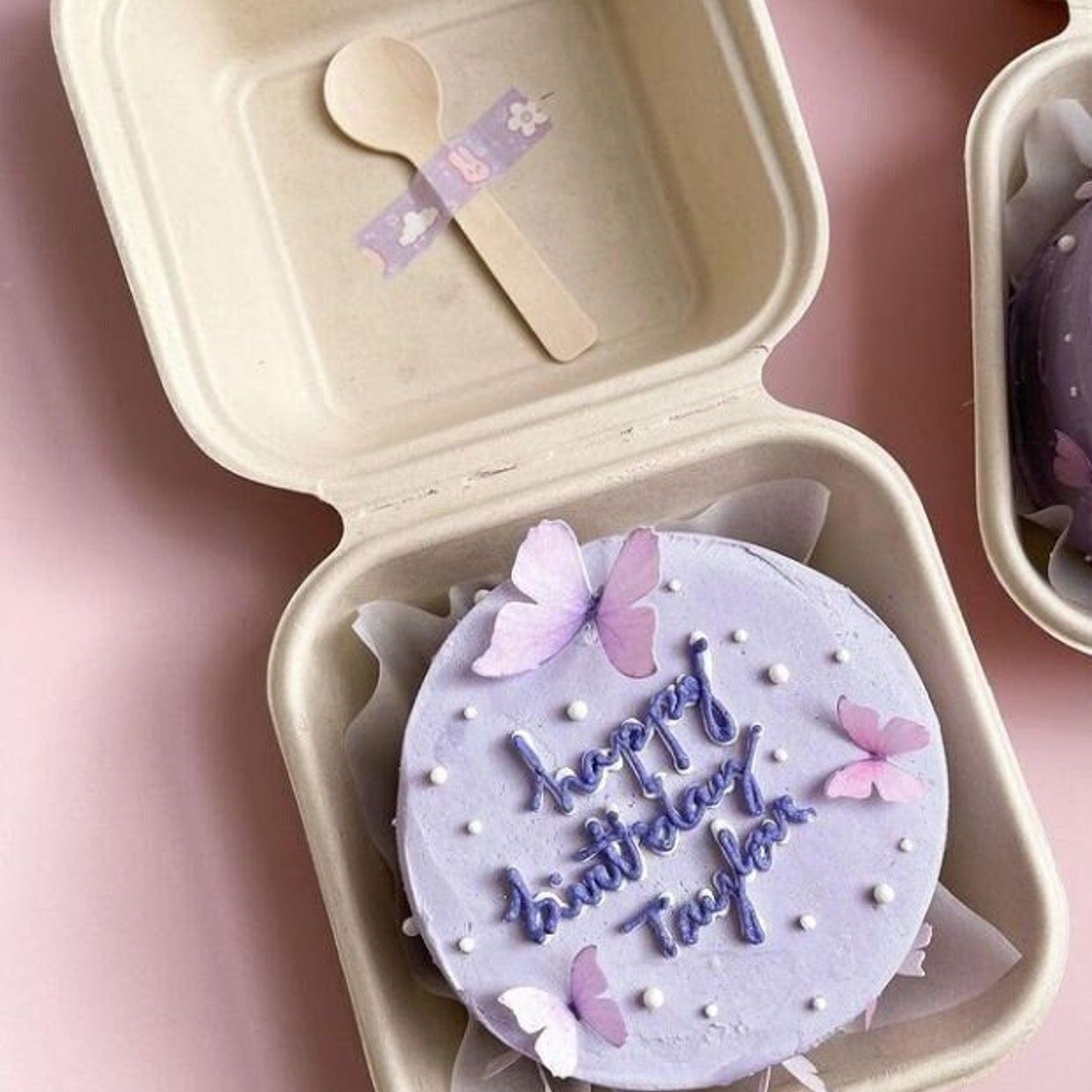 Purple birthday cake with butterfly toppers inside a kraft cake gift box and wooden spoon.