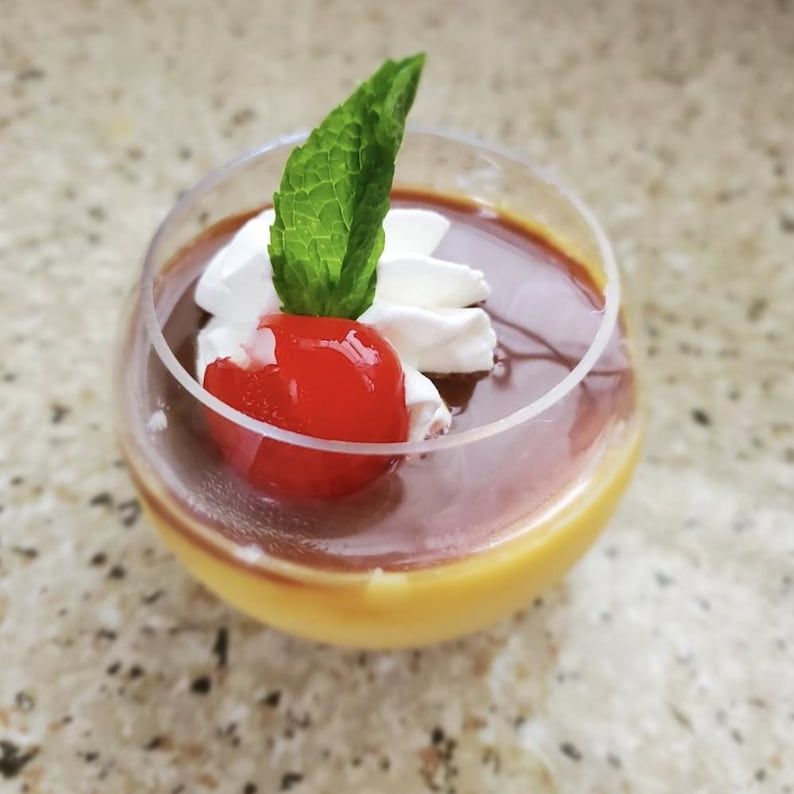 Round clear acrylic dessert bowl with layered custard, chocolate, whipped cream, and cherry