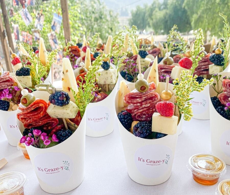White charcuterie paper cups filled with cheese, fruit, and meat appetizers on gold fancy toothpicks for party display.