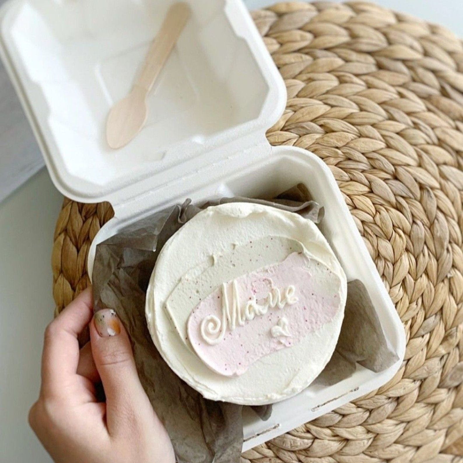 White frosted cake placed inside a kraft cake gift box with a wooden spoon.