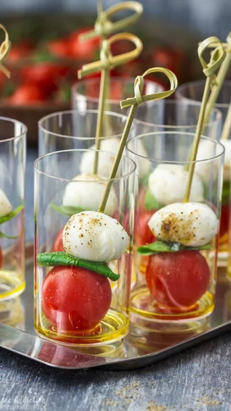 Appetizer skewers with mozzarella balls, cherry tomatoes, and basil leaves in small glasses on a tray.