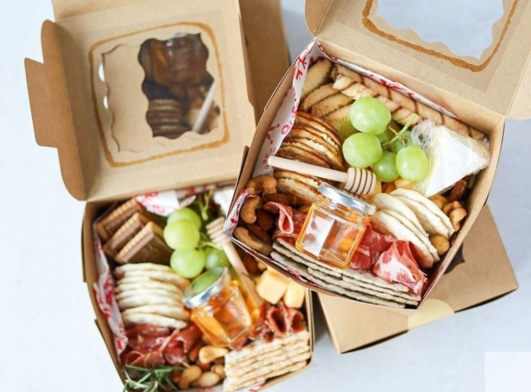 Brown square charcuterie favor boxes with clear lids filled with cheese and fruit snacks
