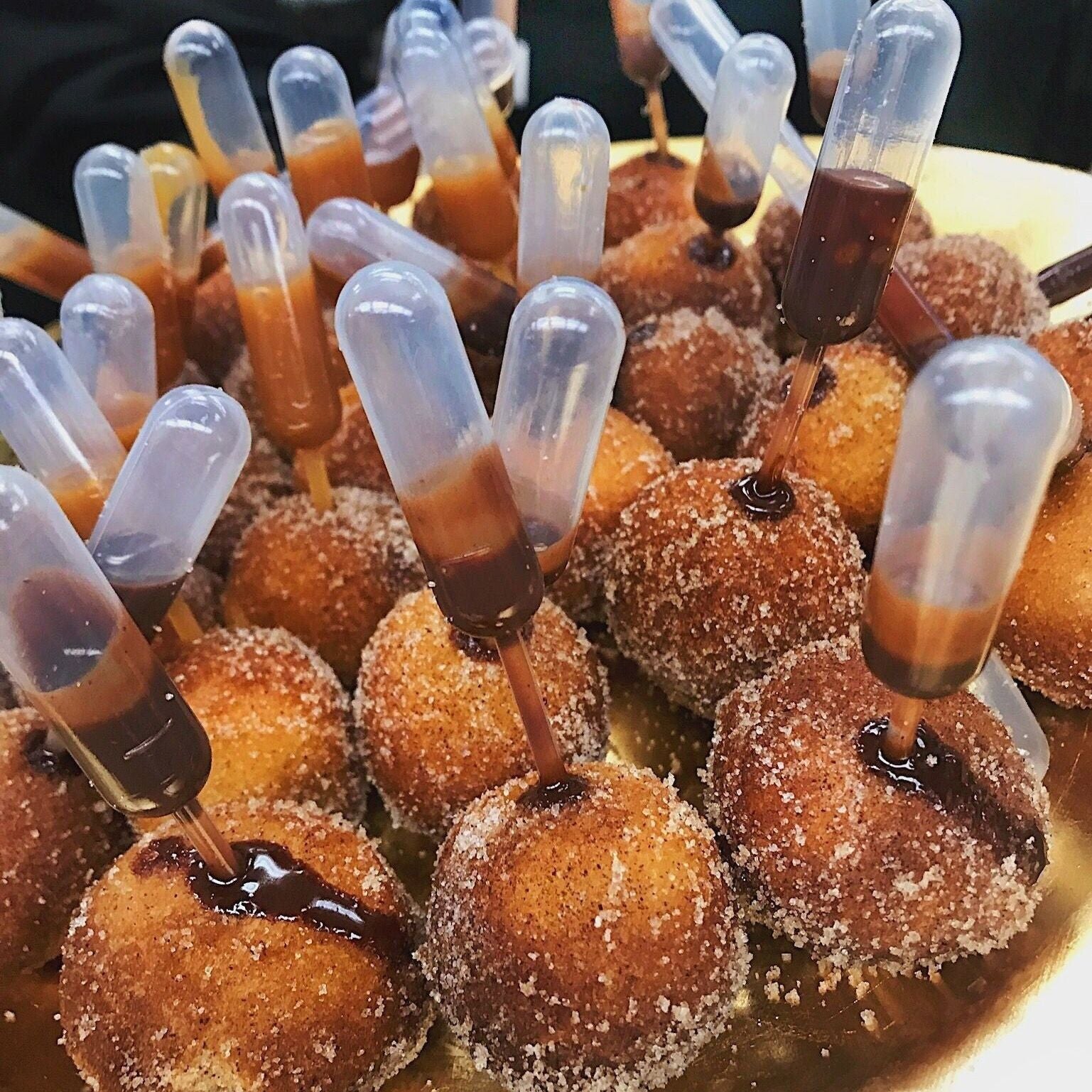 Tray of sugared donut holes each with a mini pipette filled with chocolate sauce