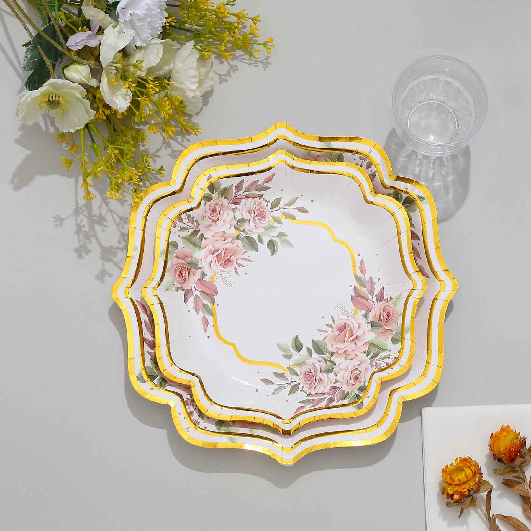 Stack of gold floral disposable scallop rim plates with glass and flowers