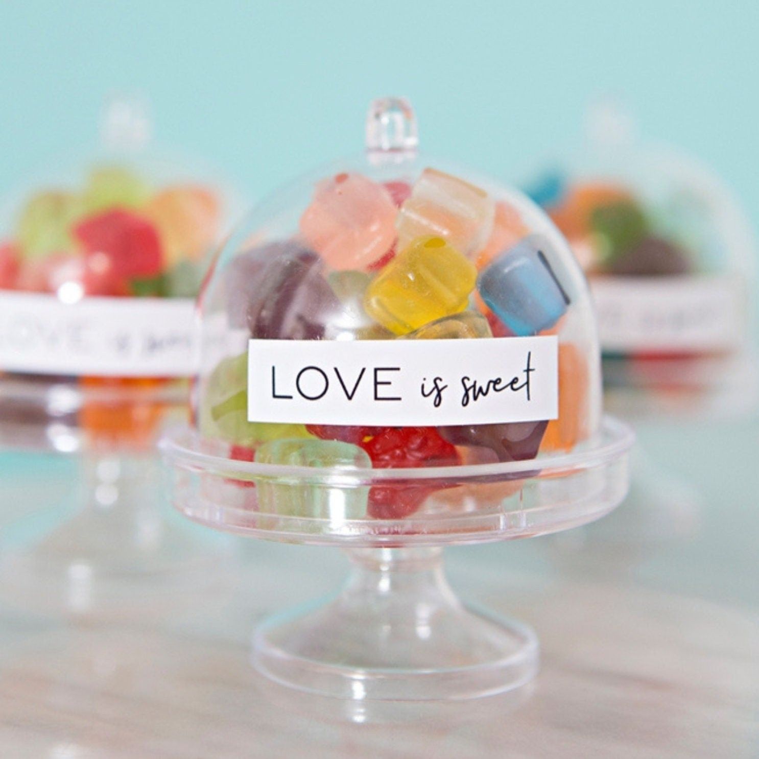 Clear mini cake dome filled with colorful candy labeled “Love is Sweet”