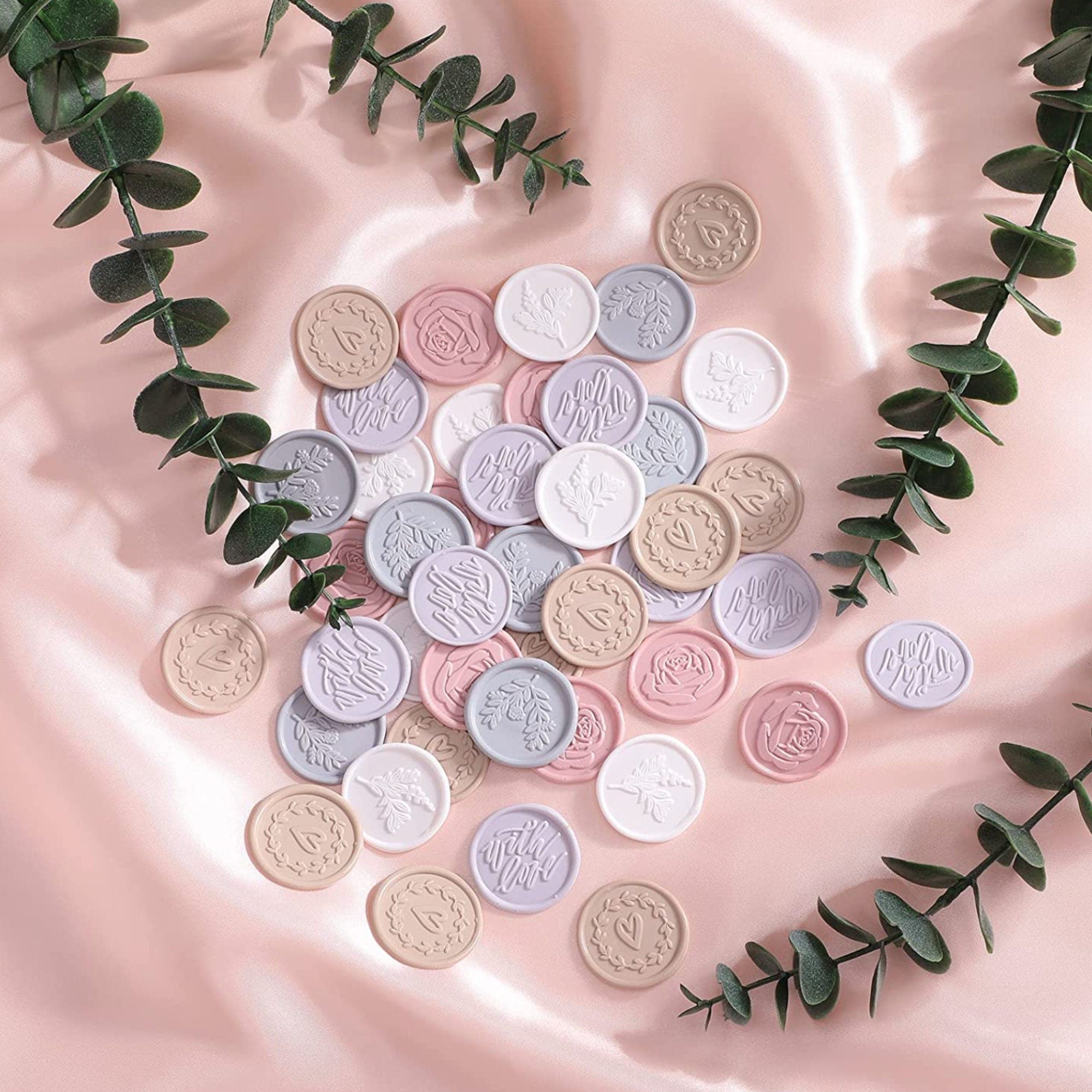 Assorted pastel 1-inch wax seal stickers displayed on blush satin with eucalyptus, showing ivory, lilac, dusty rose and beige tones.