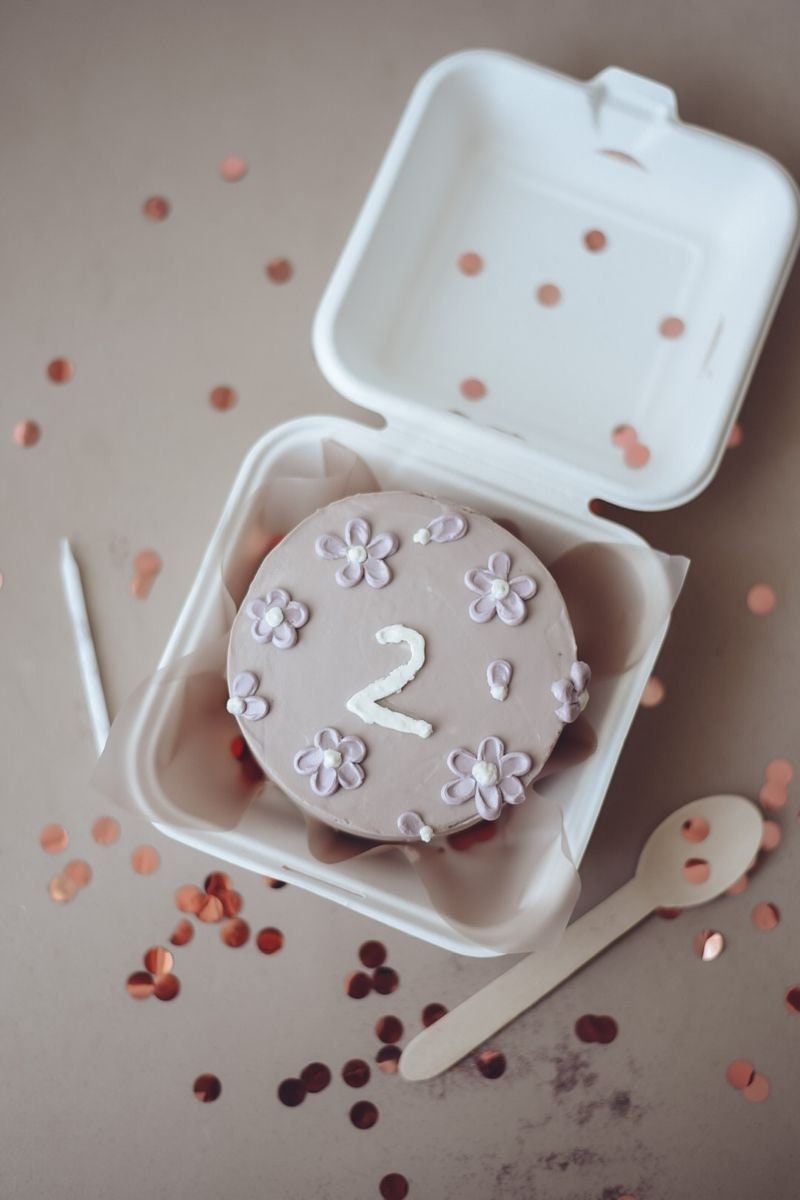 Pink floral cake displayed in a kraft cake gift box with a wooden spoon beside it.