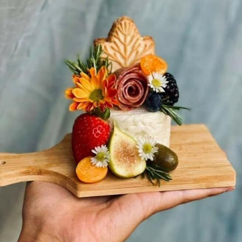 Hand holding small wooden charcuterie board styled with fruit, cheese, and flowers
