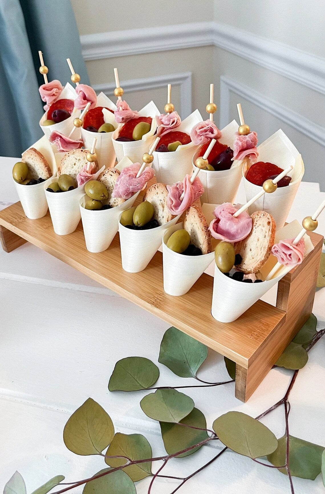Close-up of individual charcuterie cones with meat, cheese, bread, and olives, displayed on a wooden appetizer stand.